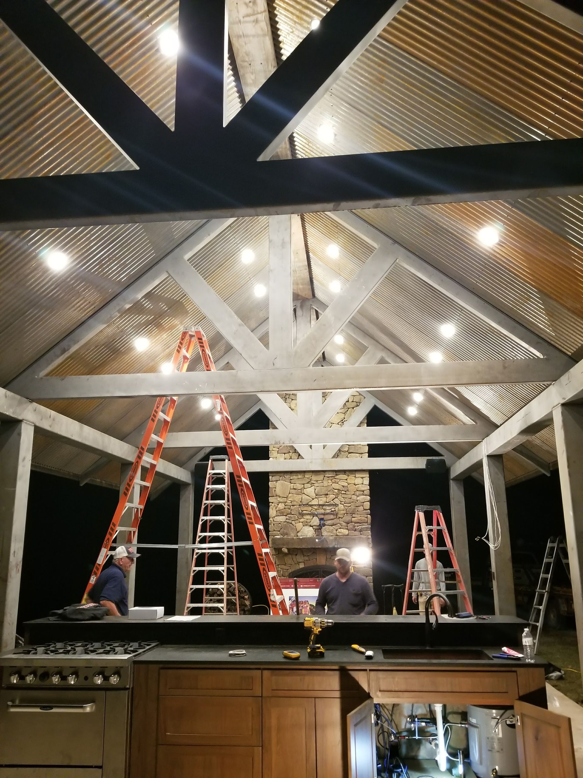 Men installing string lights on a covered patio with corrugated metal roof. Ladders, kitchen visible.