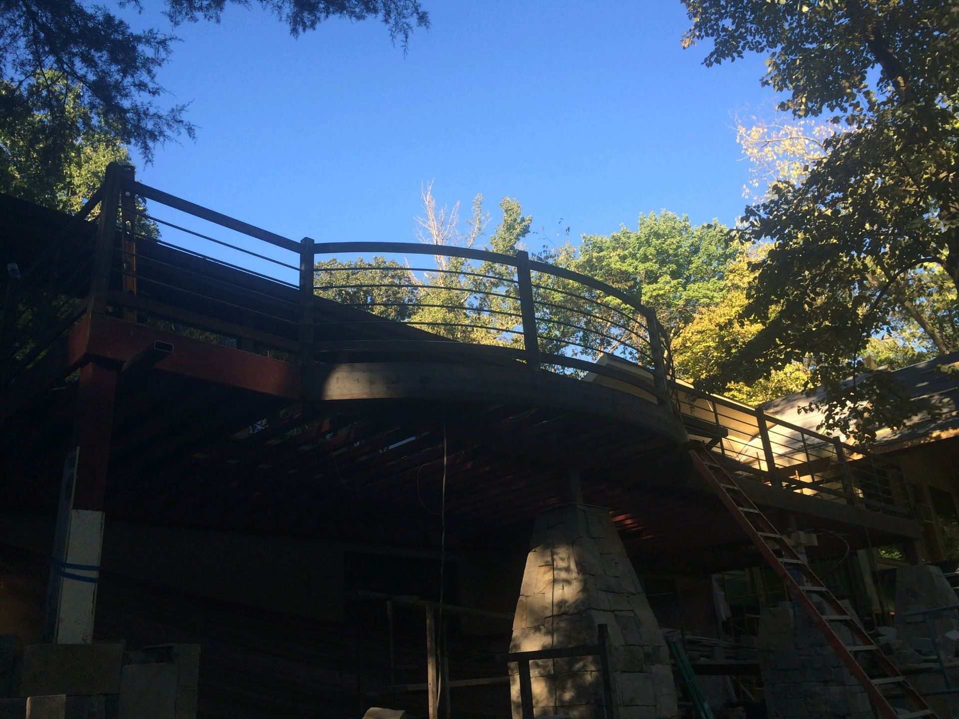 A wooden deck with a curved railing is surrounded by trees under a bright blue sky.