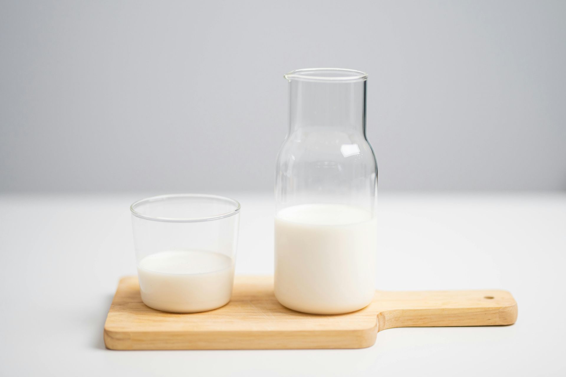 Botella de vidrio y vaso de leche sobre una bandeja de madera con fondo claro.