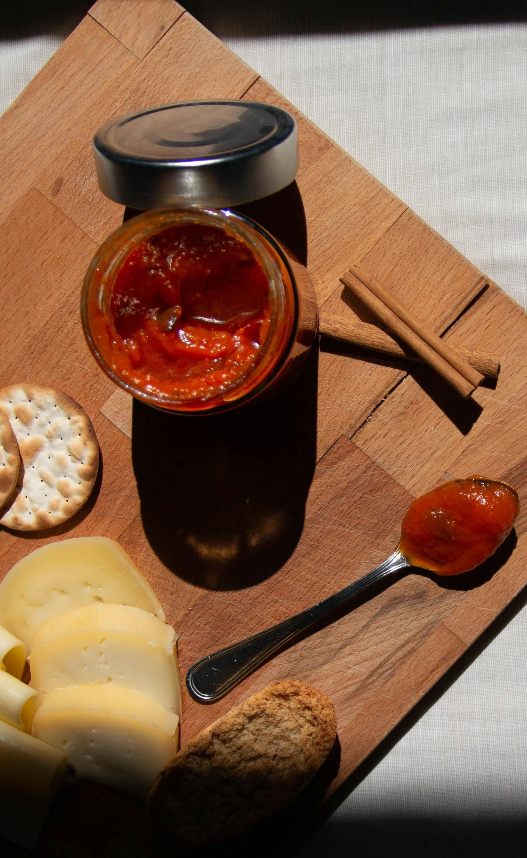 Tarro de mermelada roja sobre una tabla de madera con galletas saladas, rodajas de manzana y una cucharada de mermelada.