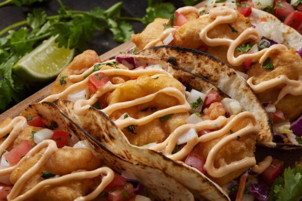Close-up of three tacos with fried shrimp, pico de gallo, sauce, and lime wedge.