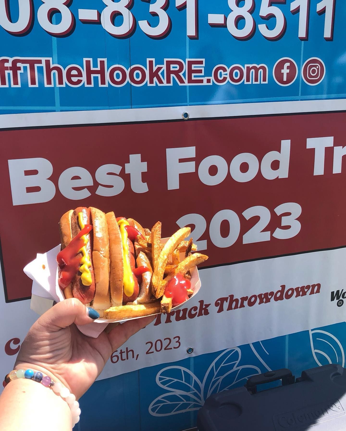 Hand holding a hot dog and fries in front of a food truck sign reading 