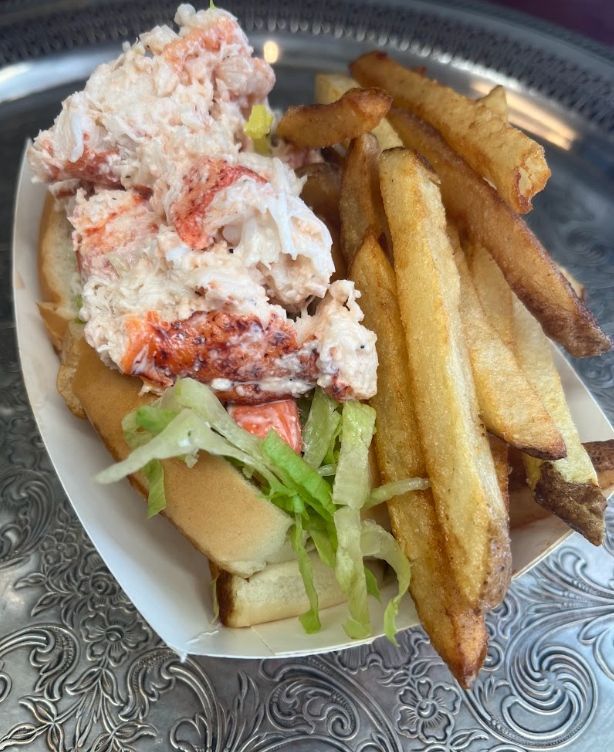 Lobster roll sandwich with fries on a silver platter. White lobster meat piled high, lettuce, and golden fries.