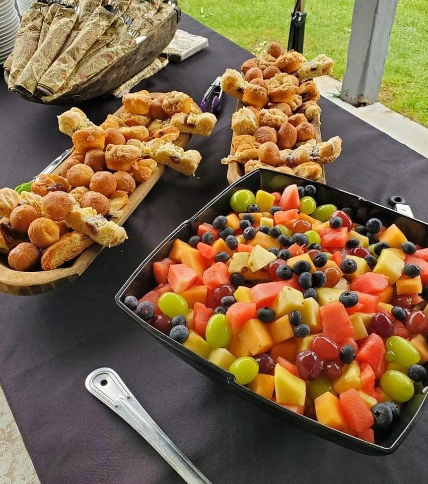 A table with fruit salad and pastries on wooden platters, set on a dark tablecloth outdoors.