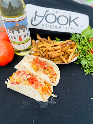 Two fish tacos with fries, a bottle of white wine, tomatoes, and cilantro, set on a black surface.