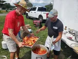Two men preparing food outdoors, one adding ingredients to a large pot, with a white truck in the background.