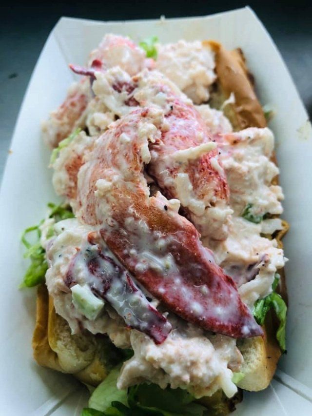 Lobster roll on a bun in a paper container. Large chunks of lobster meat and sauce atop green lettuce.