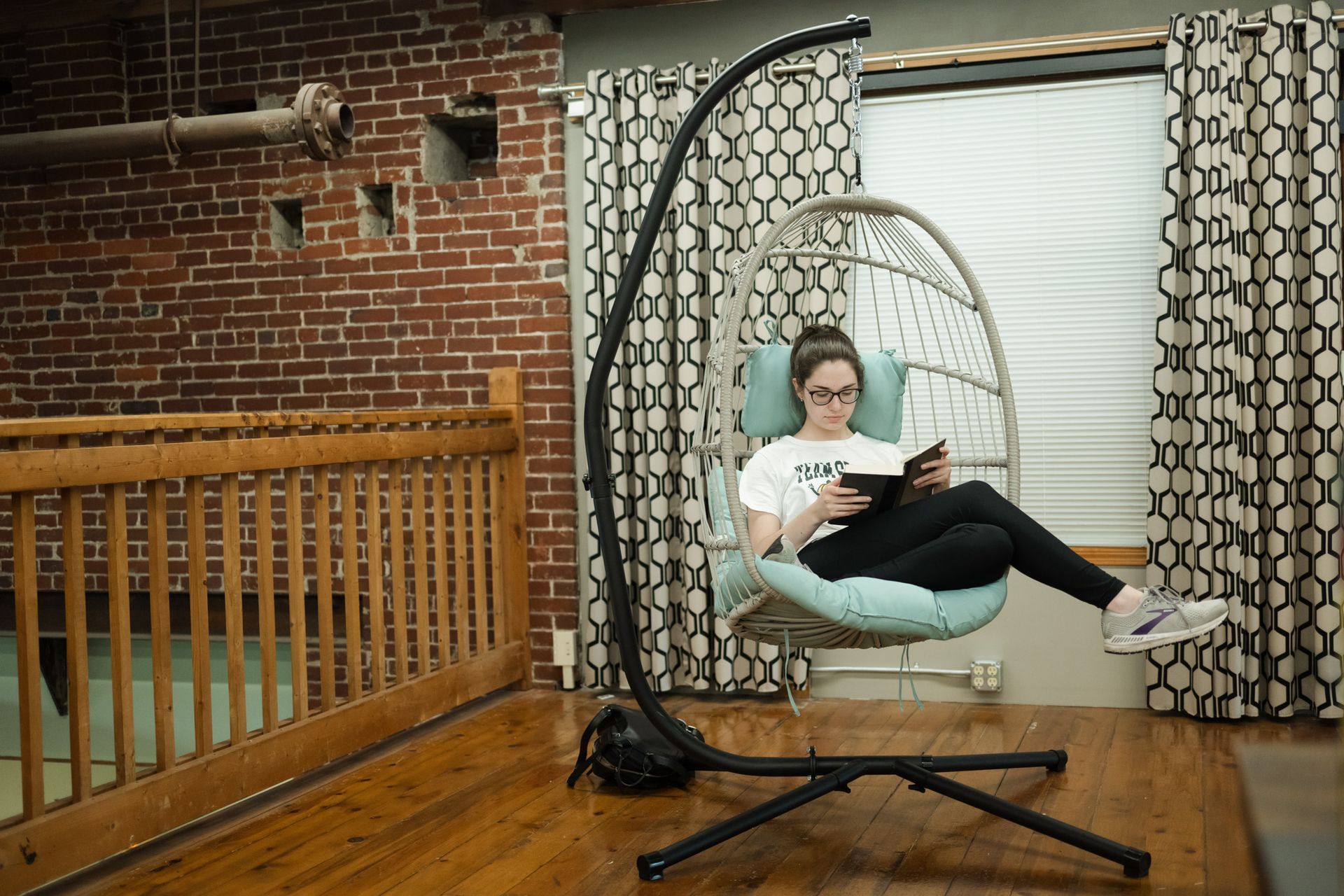 A woman is sitting in a hanging chair reading a book.