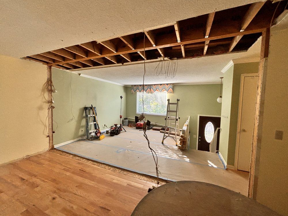 Interior undergoing renovation, showing exposed ceiling beams, opened wall, and various tools, with a living room in the background.