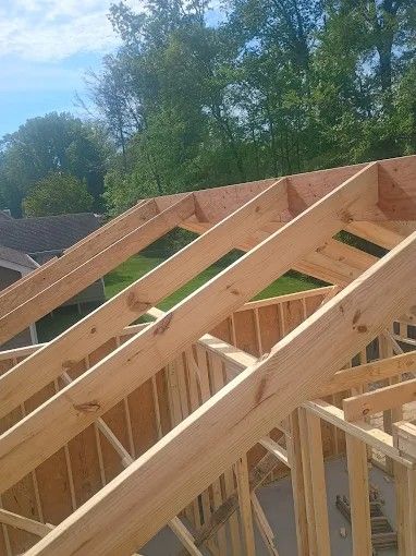 Construction framing of a house roof. Wooden rafters and sheathing are in place against a green backdrop.