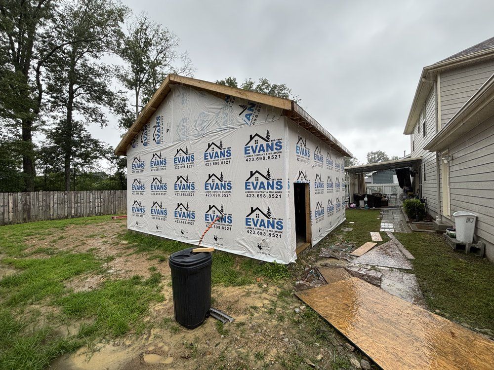 A newly constructed garage with blue-and-white wrap, doorway, and framing in a backyard, under a cloudy sky.