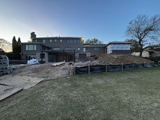 Backyard construction site with a two-story gray house, dirt pile, black barrier, and grass.