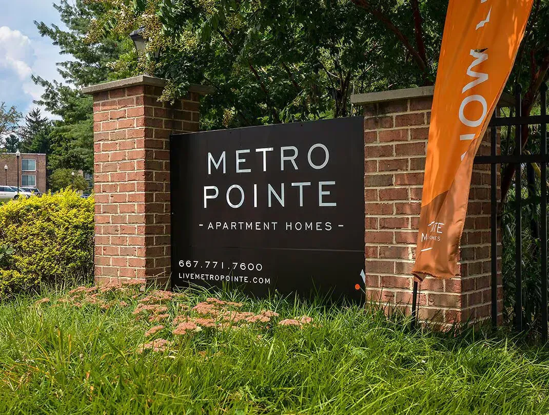 Brick entry sign reading Metro Pointe Apartment Homes with landscaping and an orange banner at Metro Pointe in Baltimore, MD.