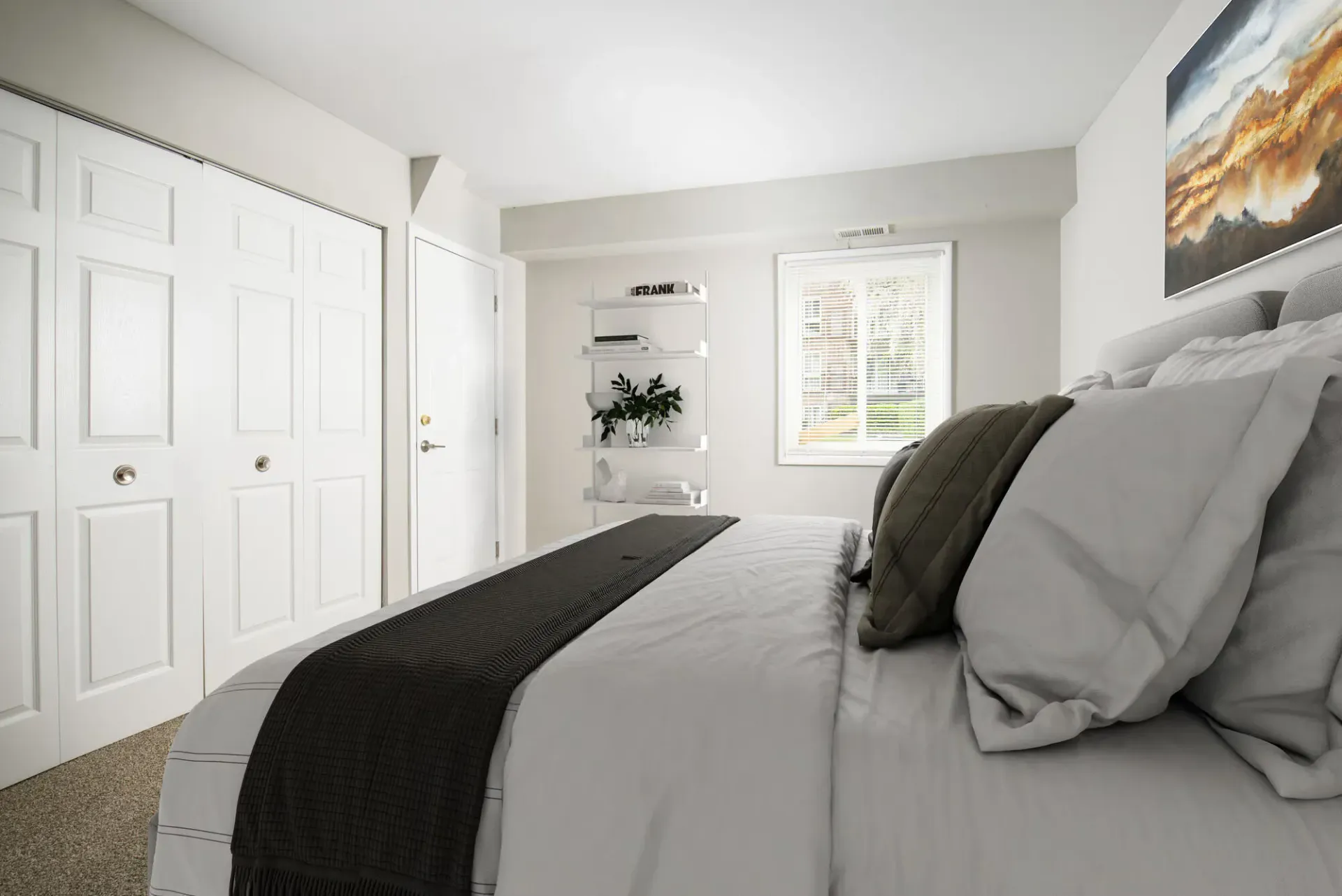 Bedroom with a neatly made bed, white walls, closet doors, and a window with a shelving unit nearby at Metro Pointe in Baltimore, MD.