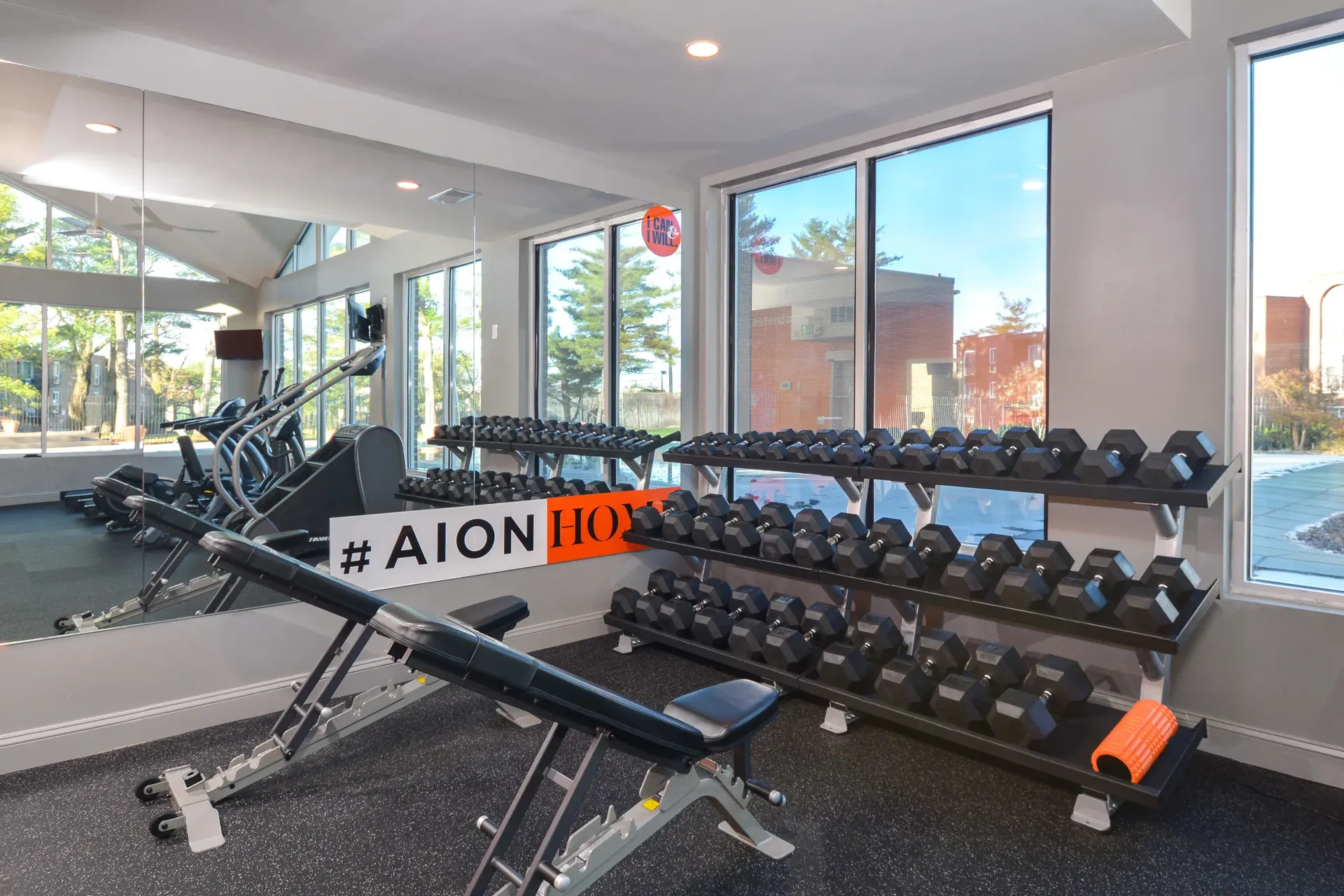 Interior community gym with dumbbells on racks, a bench press, and treadmills by large windows at Metro Pointe in Baltimore, MD.