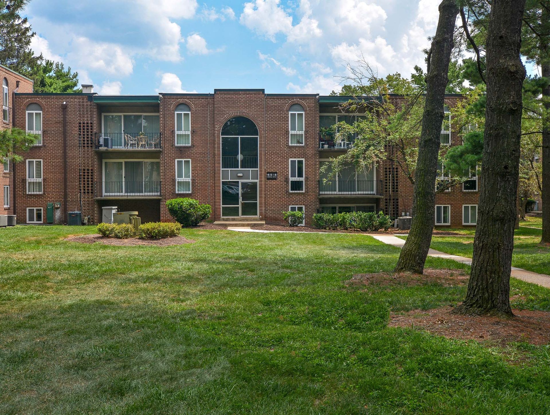 Exterior of a brick apartment building with balconies, green lawn, and large trees at Metro Pointe in Baltimore, MD.