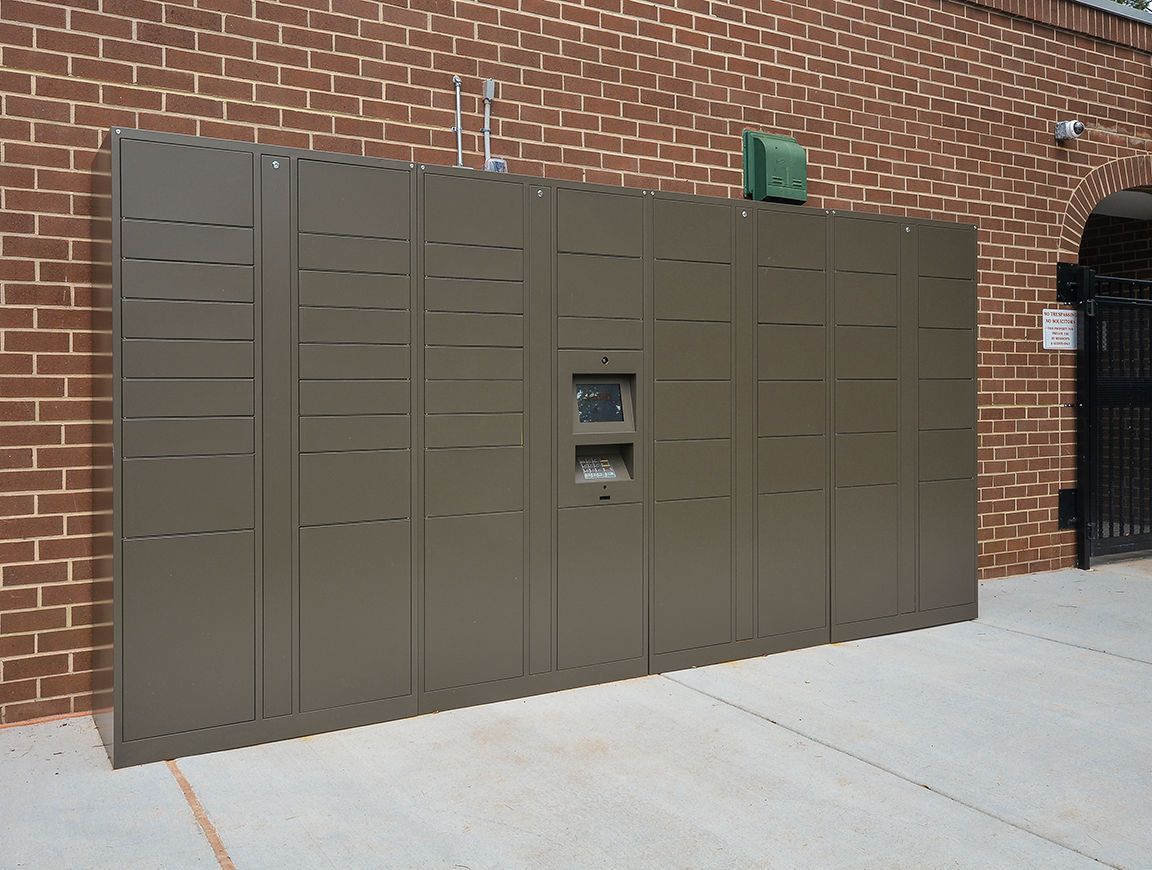 Exterior row of brown metal parcel lockers against a brick wall, with a small control panel at Metro Pointe in Baltimore, MD.