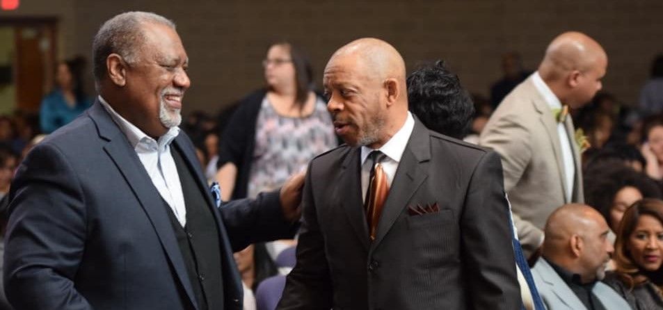 Two men in suits are shaking hands in front of a crowd.