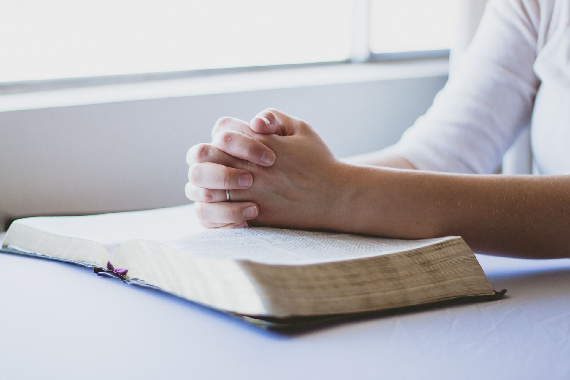A woman is sitting at a table with her hands folded over a bible.