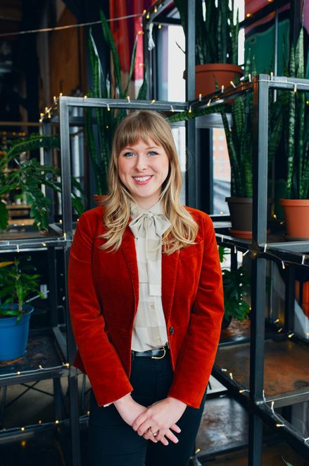 A woman in a red jacket is standing in front of a shelf with potted plants.