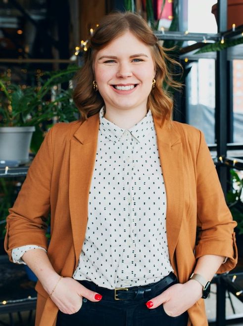 A woman in a brown jacket and polka dot shirt is standing with her hands in her pockets.