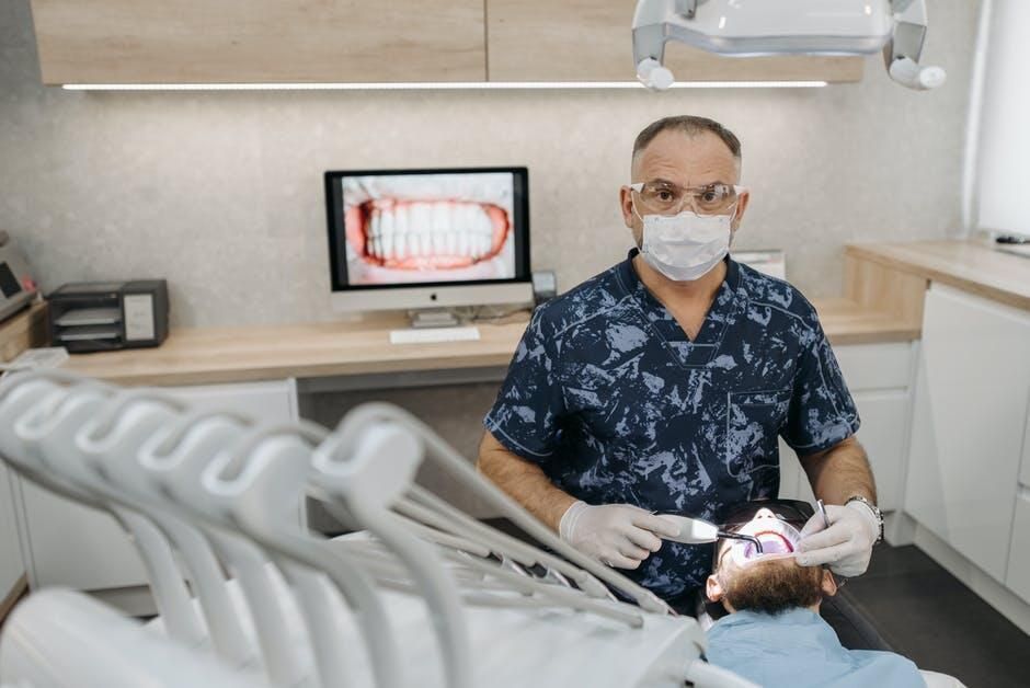 Dentist wearing mask and gloves examining a patient in a dental chair; monitor shows teeth.