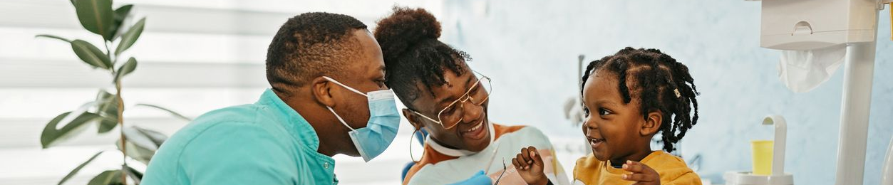 A doctor wearing a mask examines a child with the child's mother nearby.