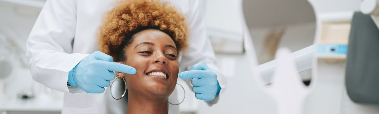 Woman smiles in a dentist's chair. She has dark hair and a red top. The setting appears to be a dental office.
