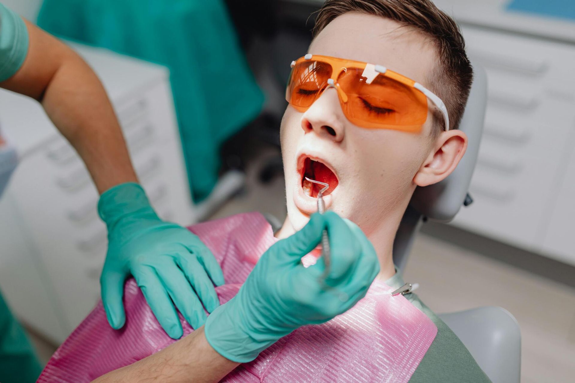 A dentist examining a patient's open mouth with tools. The patient wears orange glasses, in a dental office.