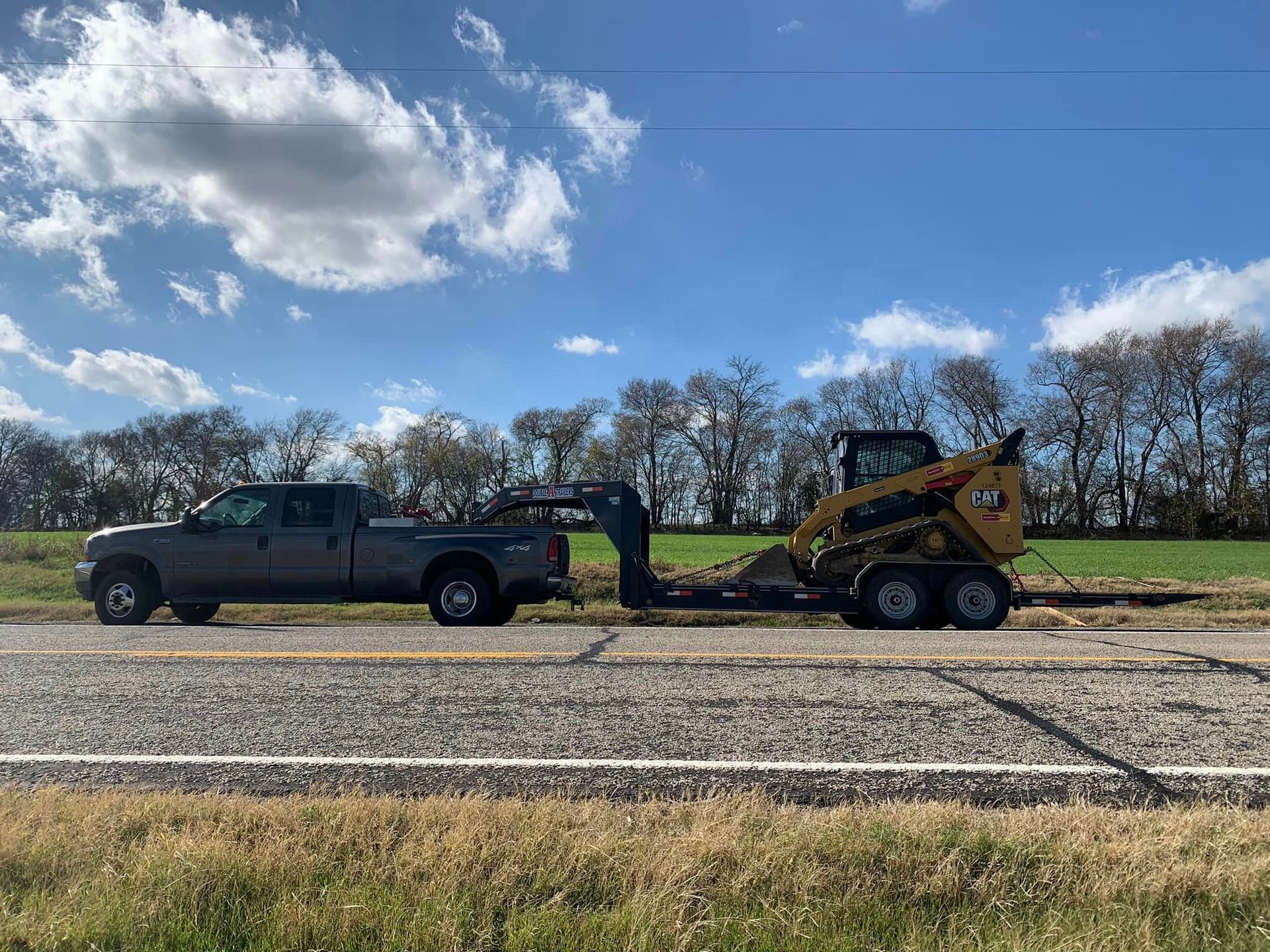 Grey pickup truck towing a construction trailer with a yellow skid steer on a roadside under a blue sky.