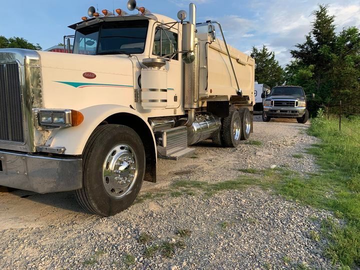 Cream-colored Peterbilt dump truck on a gravel road. A pickup truck is parked behind it, trees in the background.