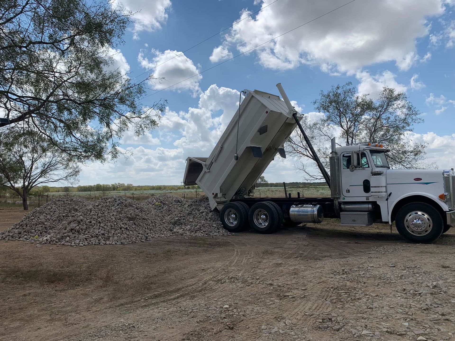 Dump truck unloading gravel onto a pile in a field under a cloudy sky.