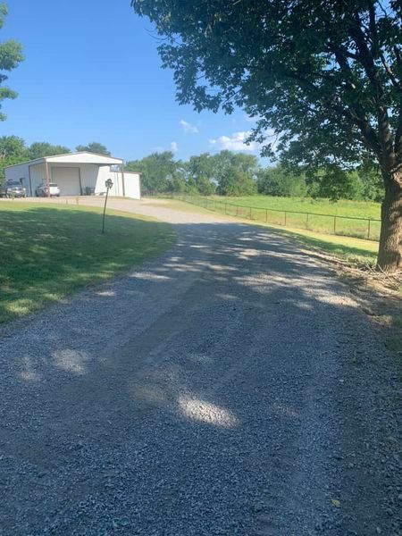 A gravel driveway leads to a white building under a bright blue sky, bordered by a tree and green grass.