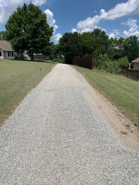 A gravel driveway leads toward trees and a house on a sunny day.