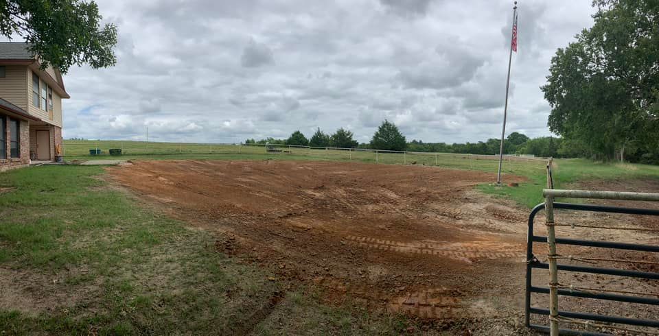 Cleared dirt lot next to a house and gated entrance; cloudy sky, trees in distance.