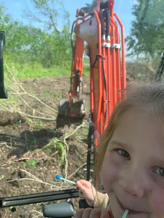 A girl smiles, holding a straw, in a tractor cab; orange excavator works in background.