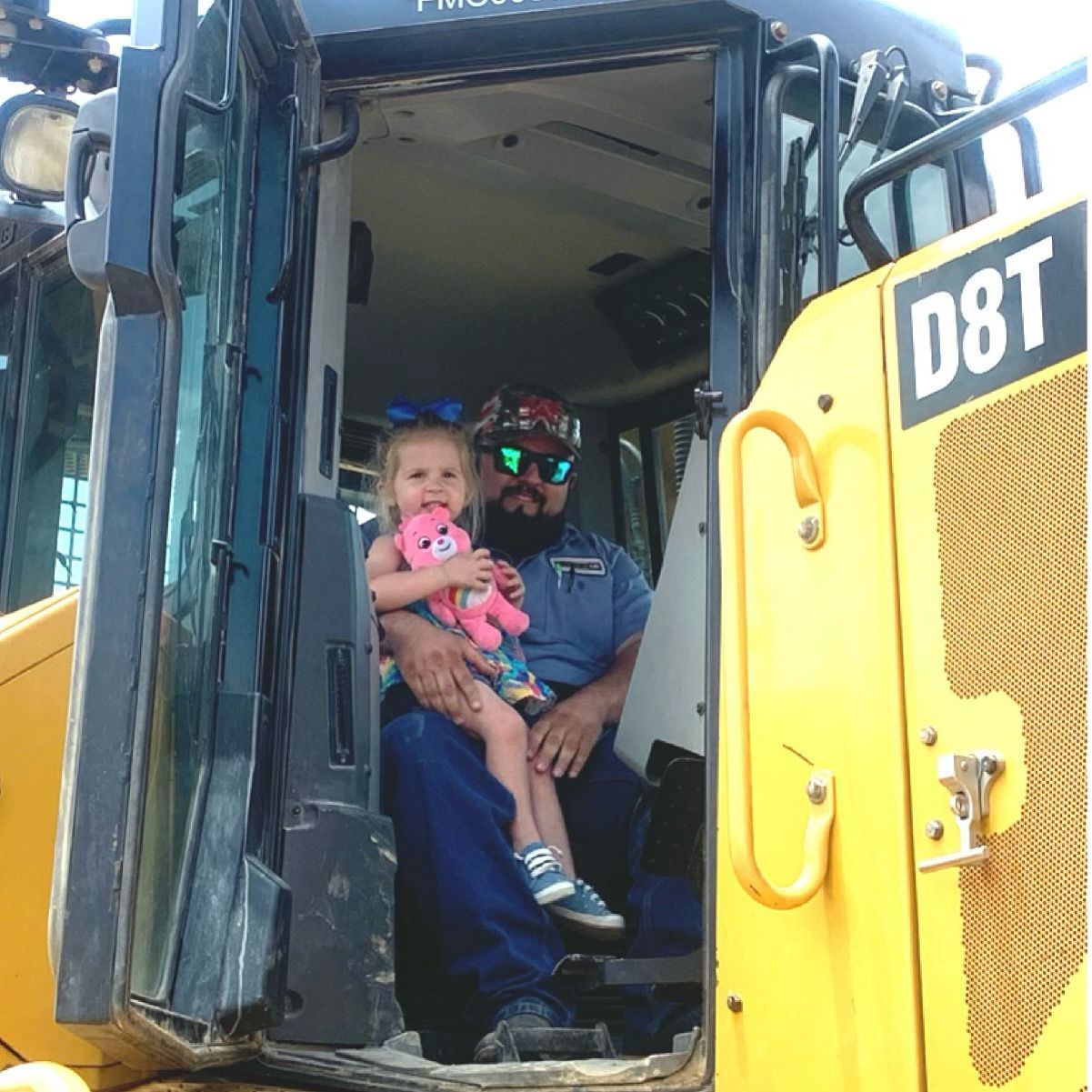 Man with a beard and young girl in a bulldozer, the man holding the child.