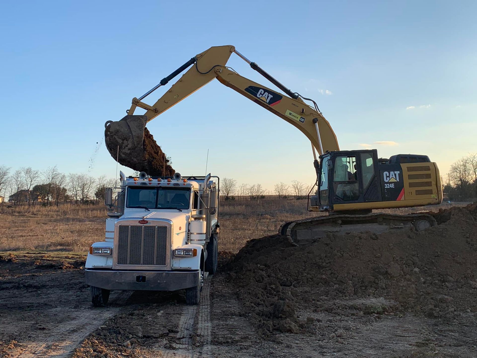 An excavator loads a white dump truck with dirt on a construction site under a blue sky.