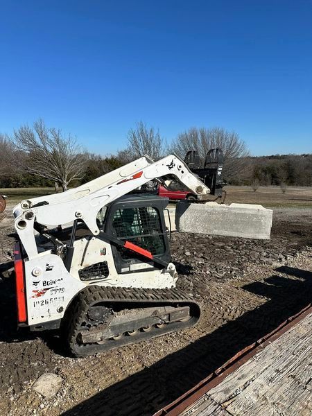 A Bobcat skid steer with a lifting attachment carrying a large concrete block in an outdoor setting.