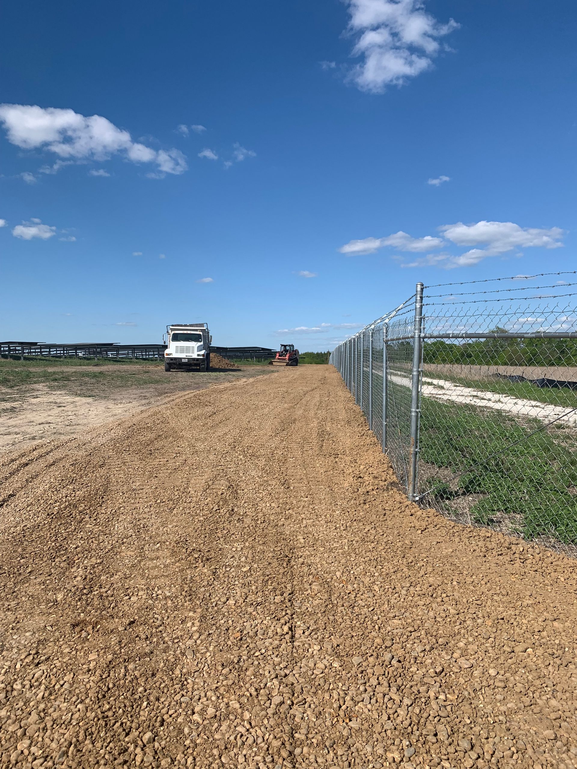A gravel path along a metal fence, with trucks and machinery on a sunny day.