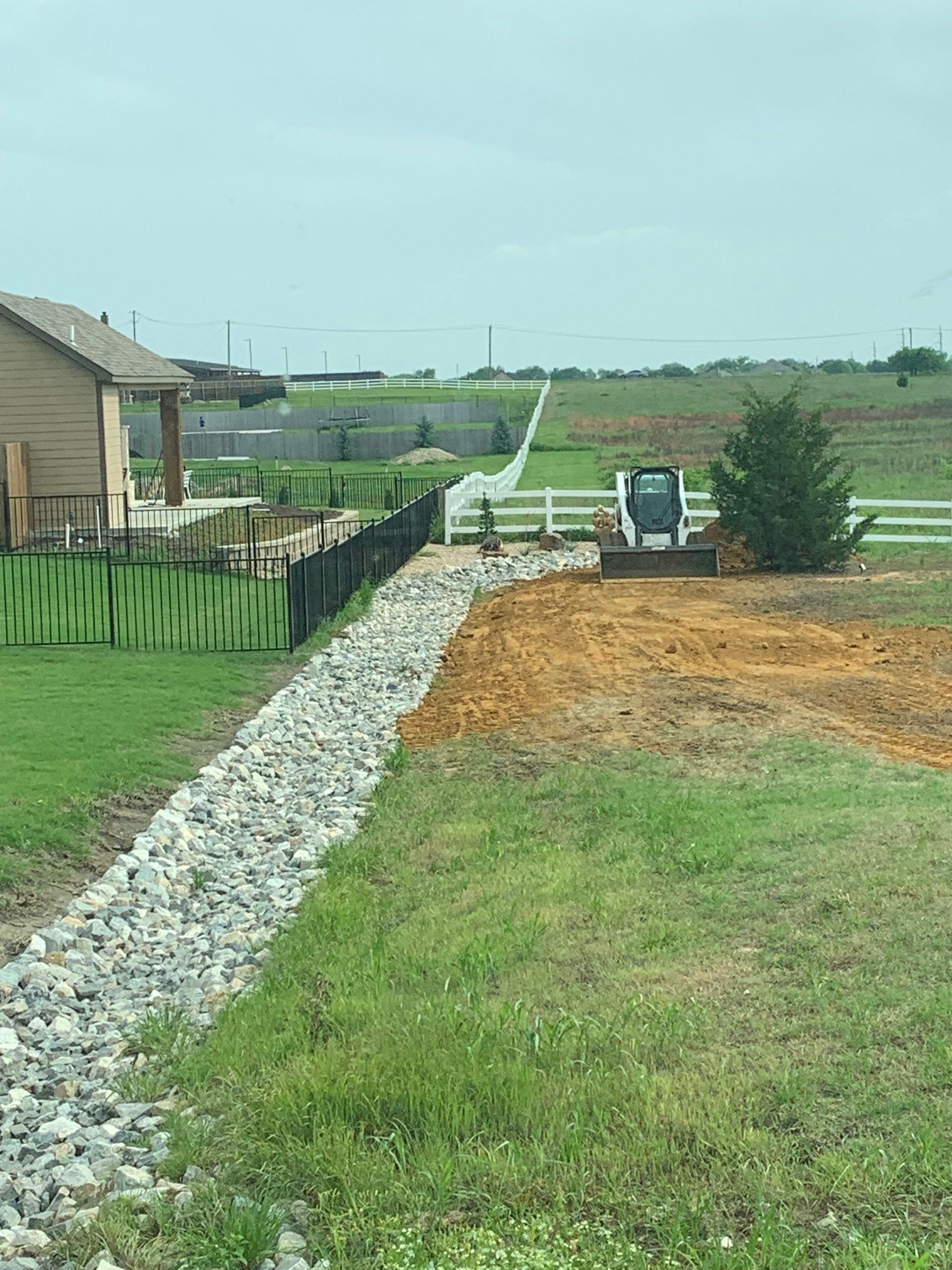 A rock-lined drainage ditch beside a house, with a white fence and a small bulldozer in the background.