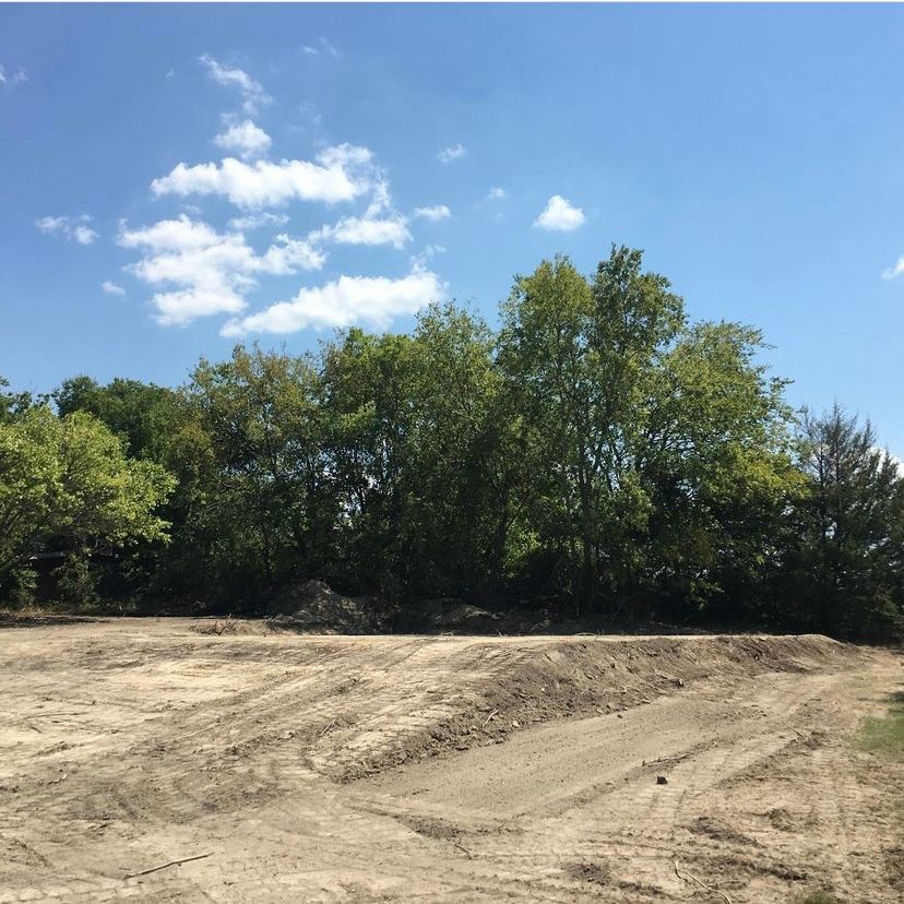 Cleared land with dirt and tire tracks in front of trees under a blue sky with clouds.
