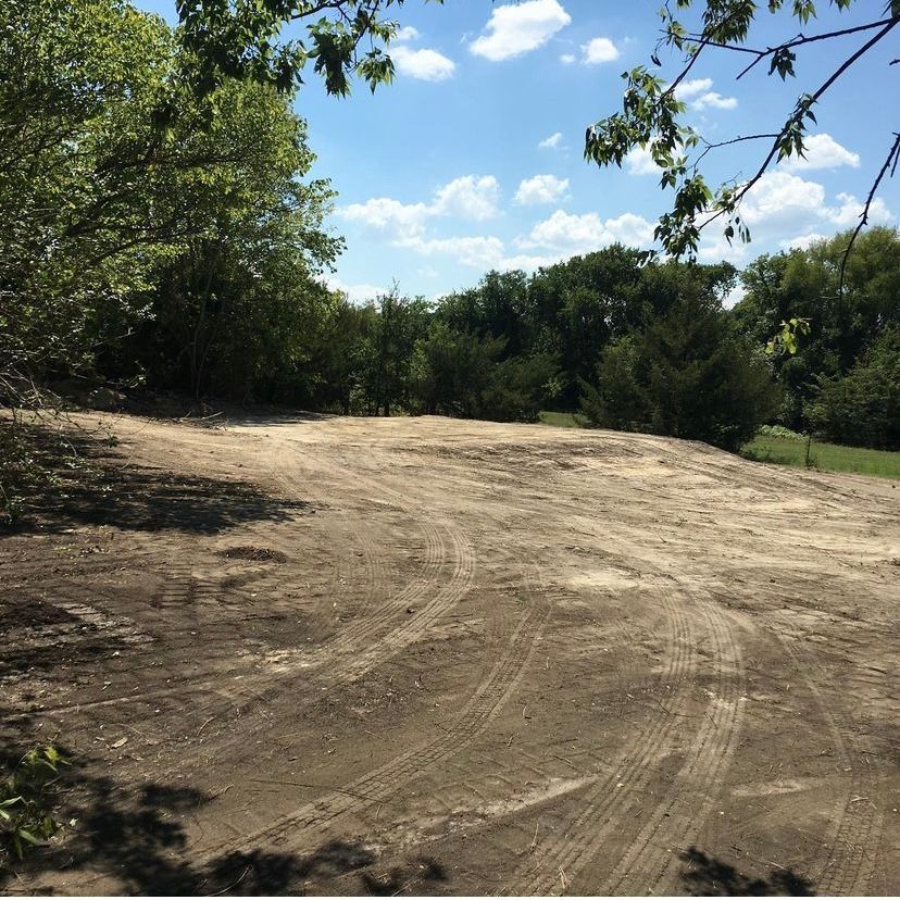Dirt clearing with tire tracks, surrounded by trees, under a blue sky.