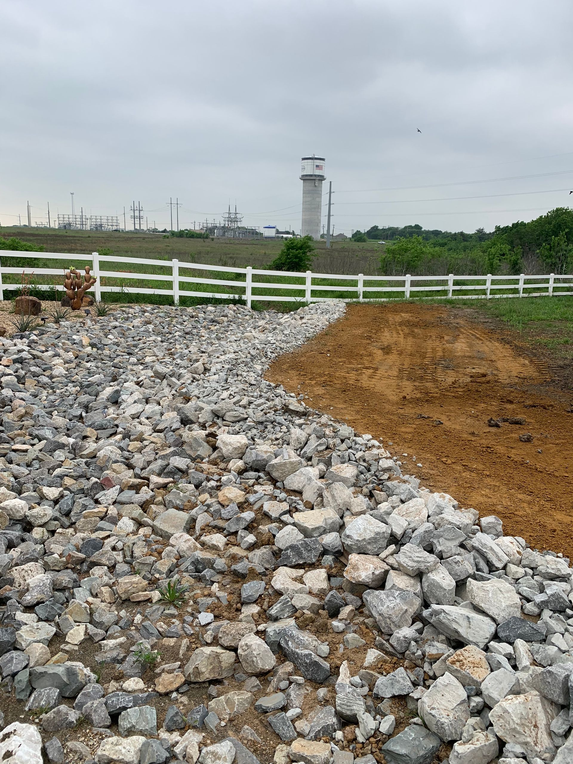 Gravel and dirt pathway next to white fence with airport tower in the distance under cloudy sky.