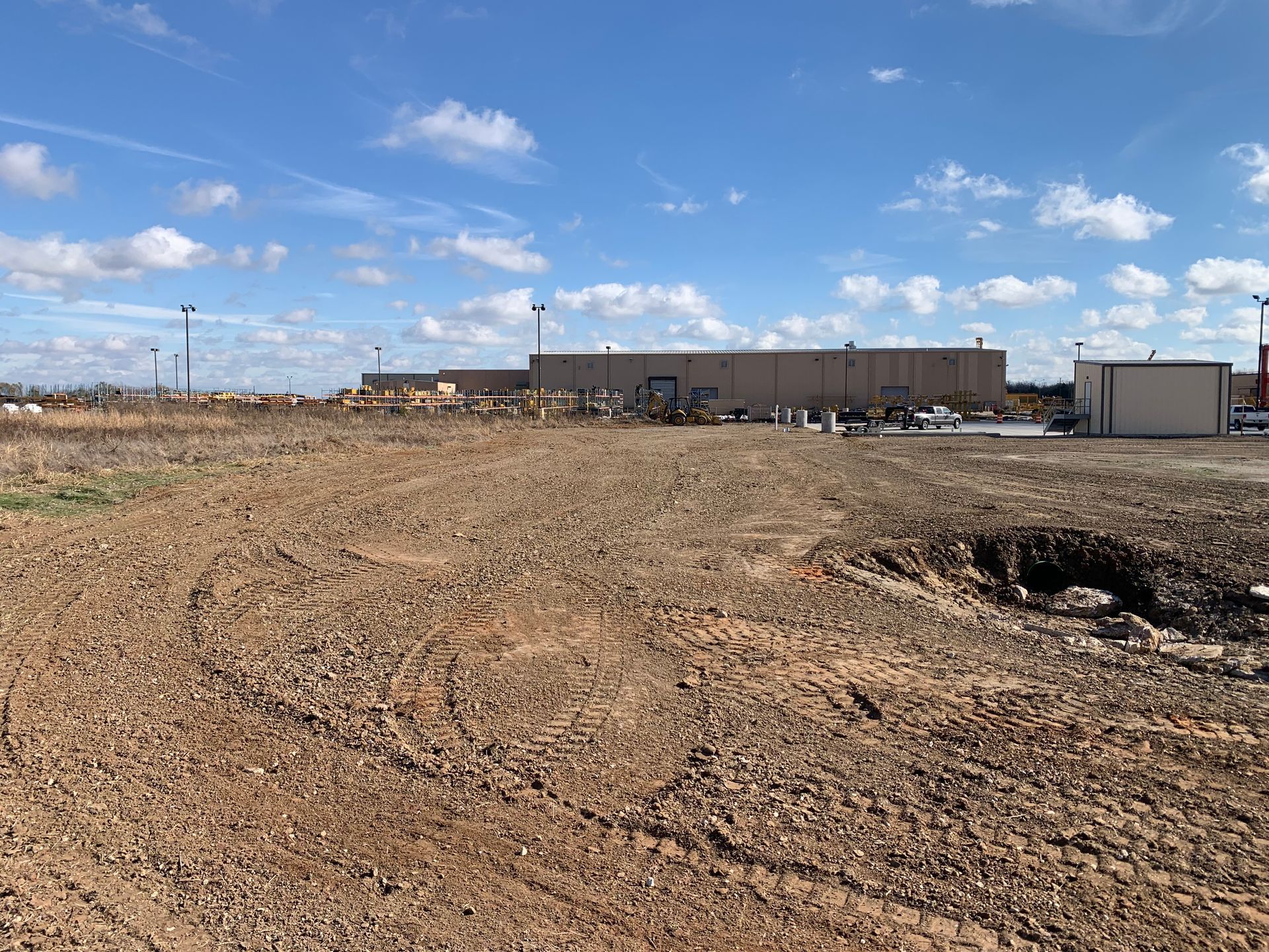 A vacant lot with gravel and dirt in the foreground, a low building in the background, under a blue sky with clouds.