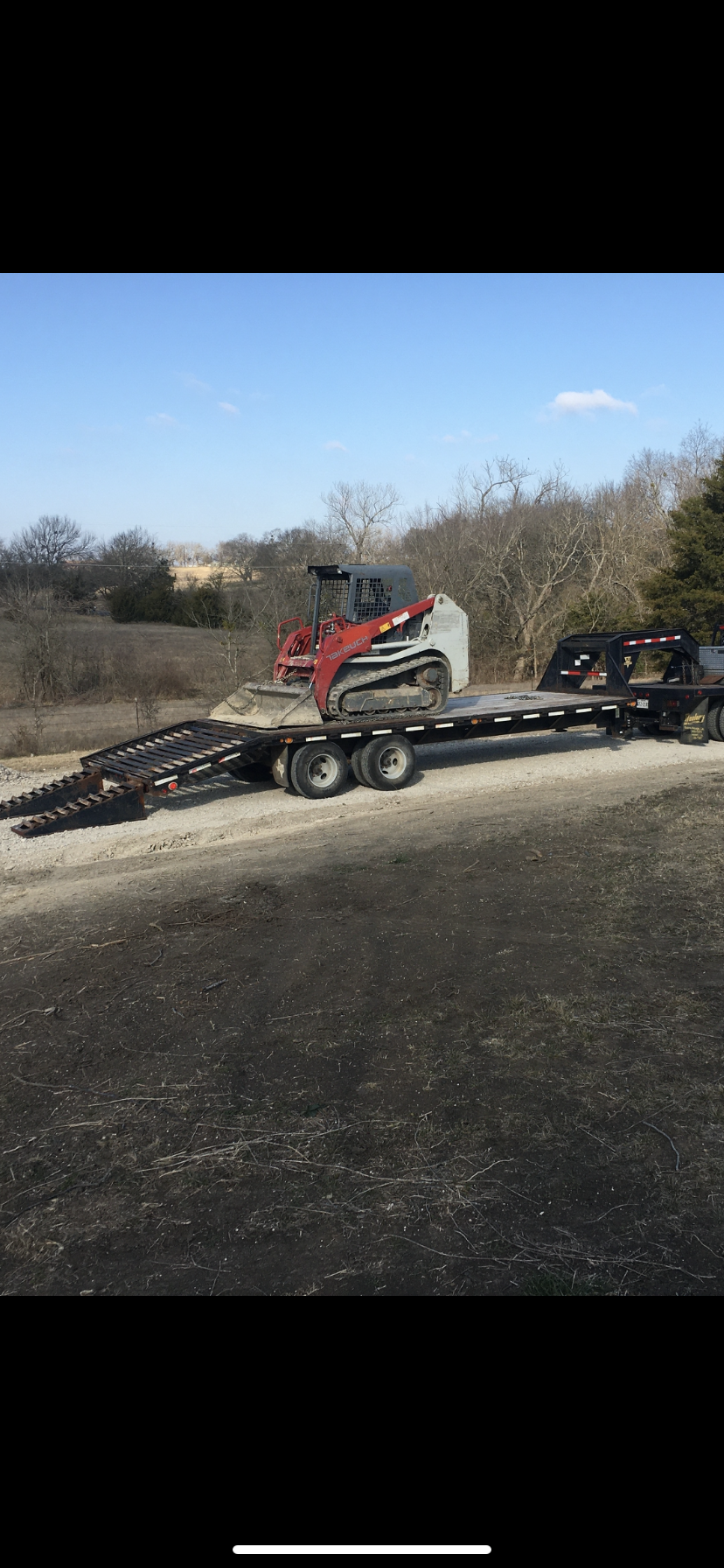 Red skid steer on a flatbed trailer, parked on an asphalt road, trees and blue sky background.