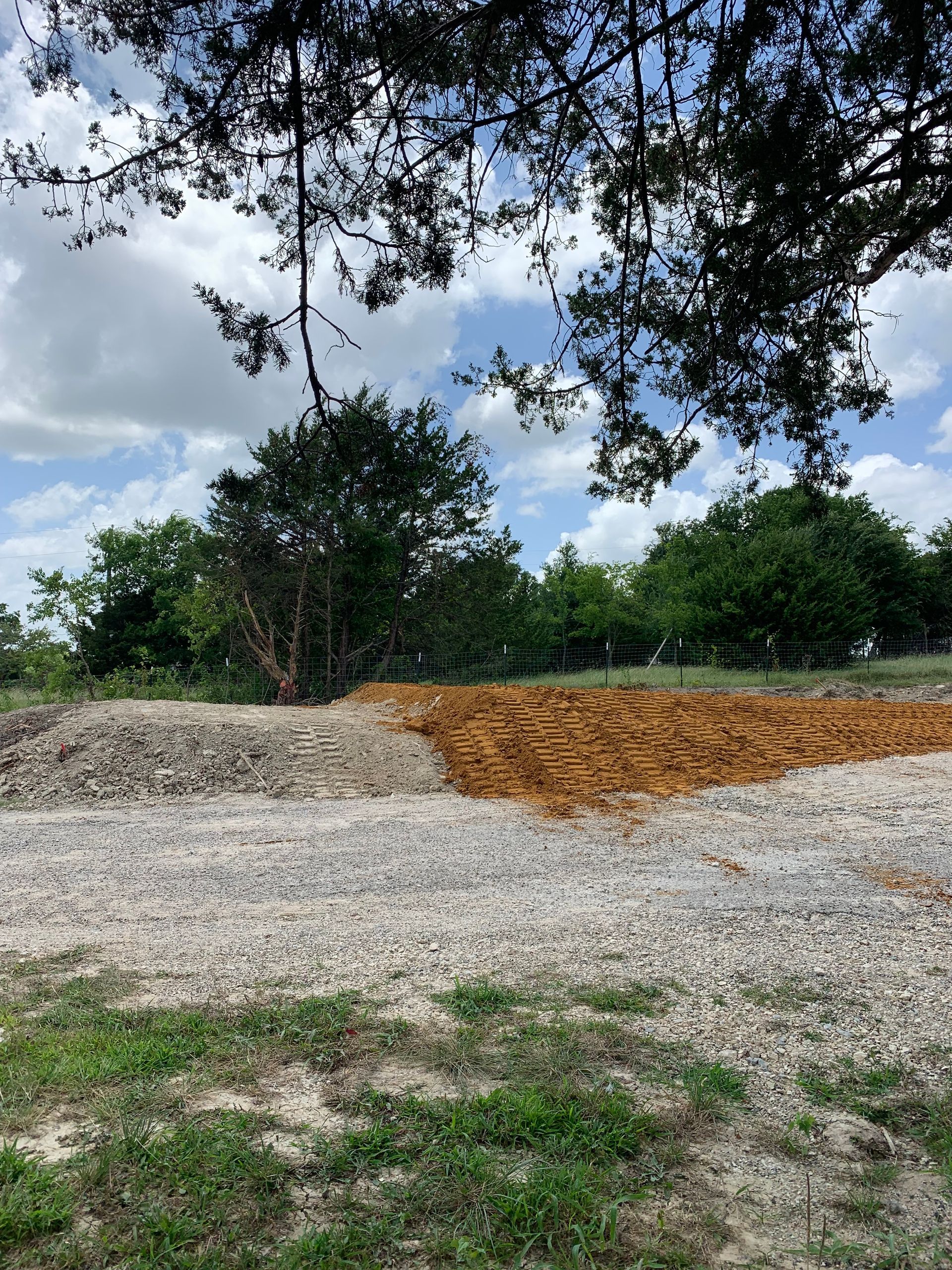 Gravel piles in a grassy field, brown and gray, under trees and a cloudy blue sky.