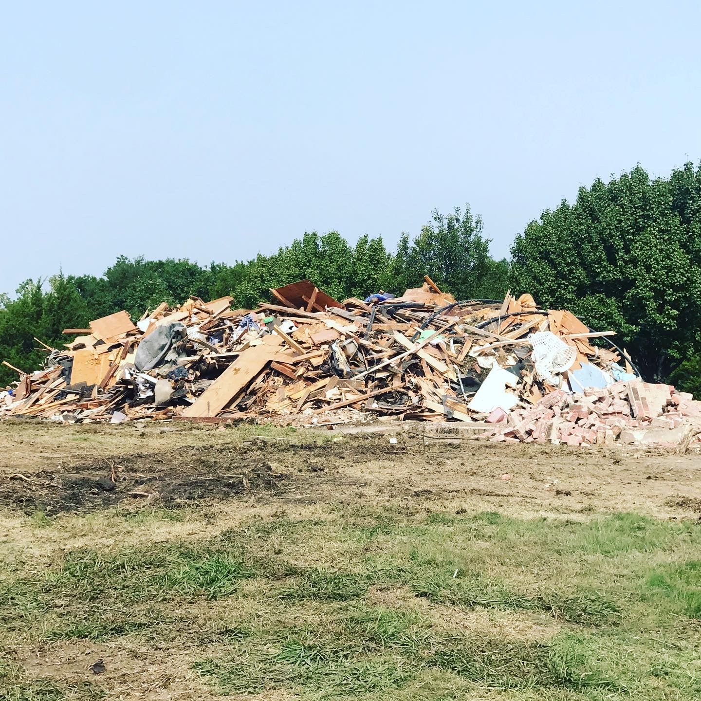 Pile of demolition debris in a grassy field with trees in the background under a clear sky.