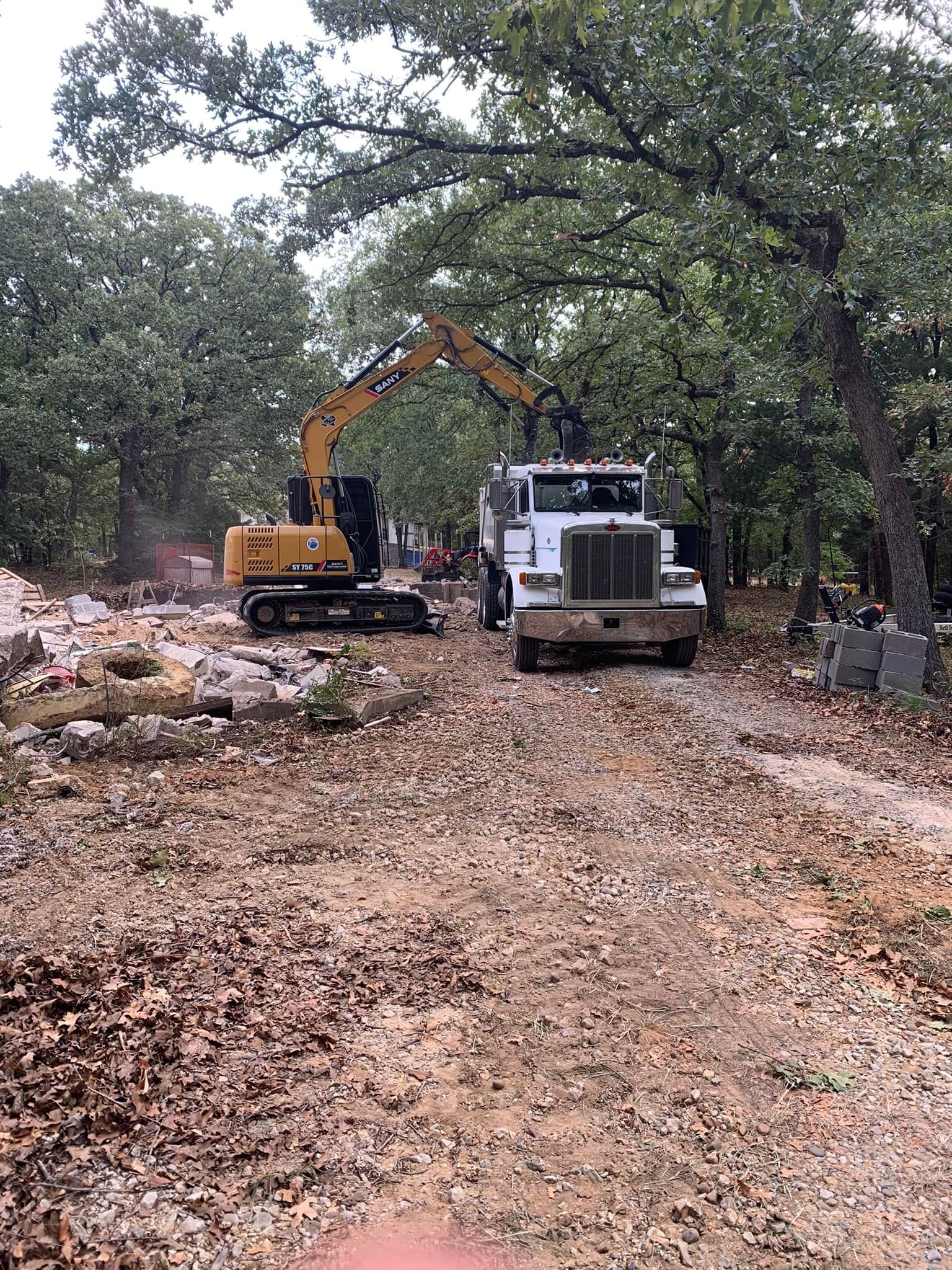 Yellow excavator loading rocks onto a white truck on a dirt road in a wooded area.