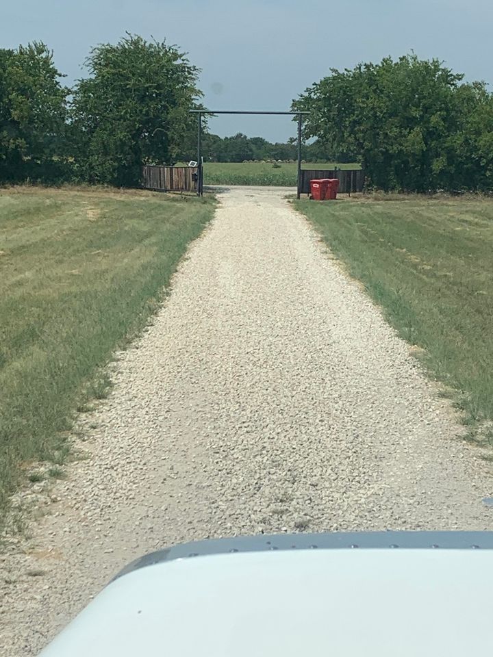 Gravel driveway leading to gate with trees on either side. Green grass and blue sky.